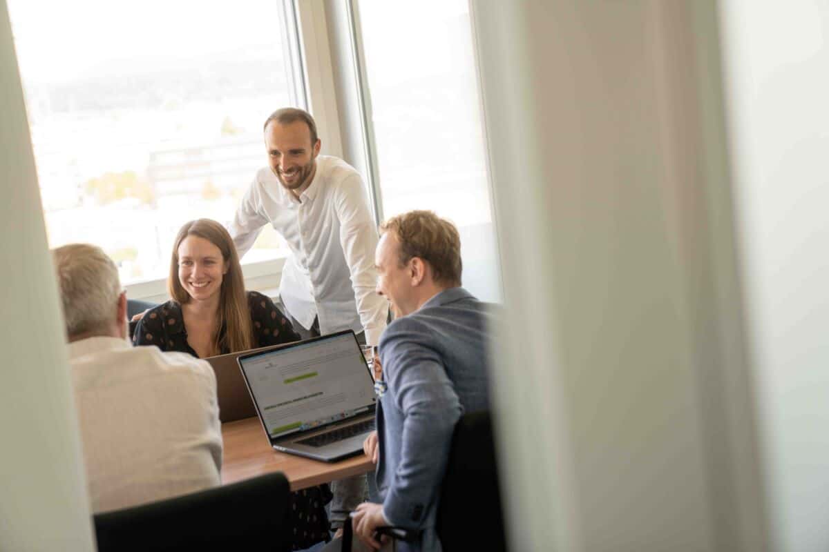 Alena, Dario, Robin und Bruno sitzen in einem Meetingraum am Tisch und arbeiten am Laptop und gleichzeitig führen sie ein Gespräch.