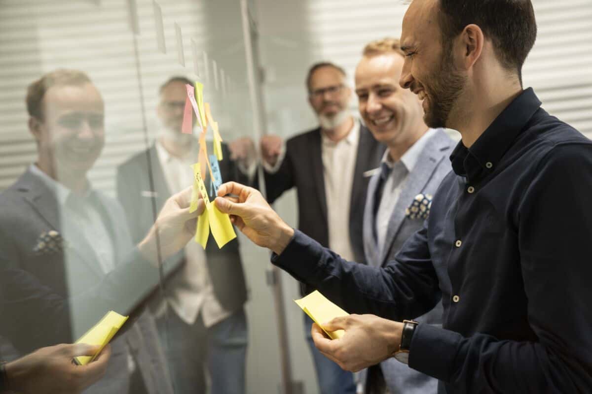 Dario, Robin und Bruno kleben Post-It auf die Glaswand und lachen dabei.
