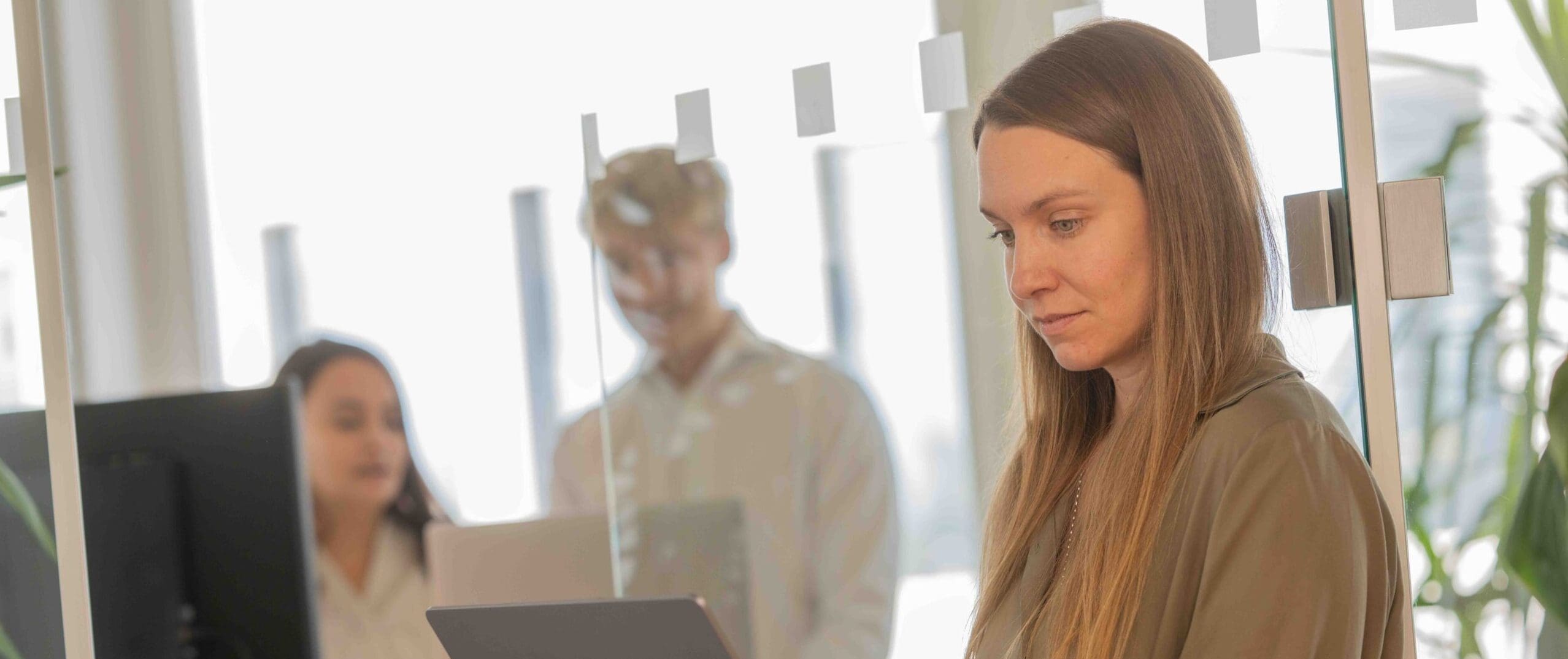 Alena schaut auf ihren Laptop, während Melanie und Sandro im Hintergrund in einer Besprechung sind.